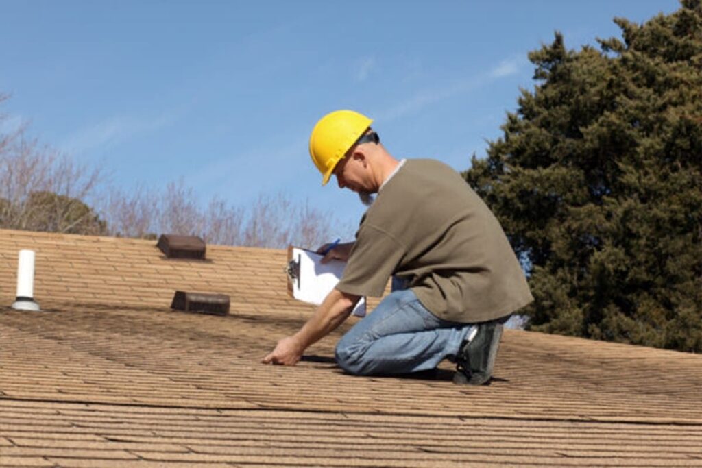 Roofing inspector examining shingles on a residential roof with a clipboard and yellow safety helmet.